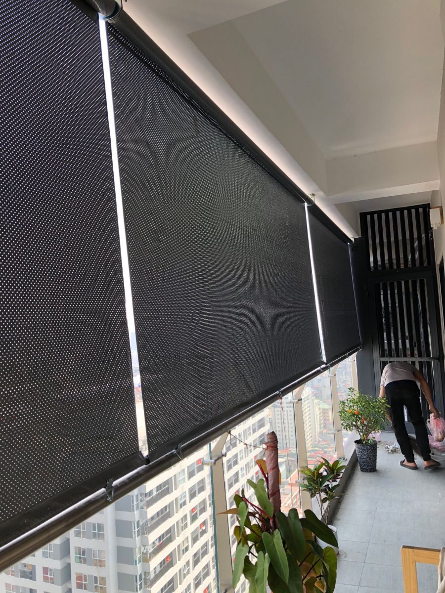 Rèm lưới cuốn màu tối che một phần ban công kính, cạnh các chậu cây.
